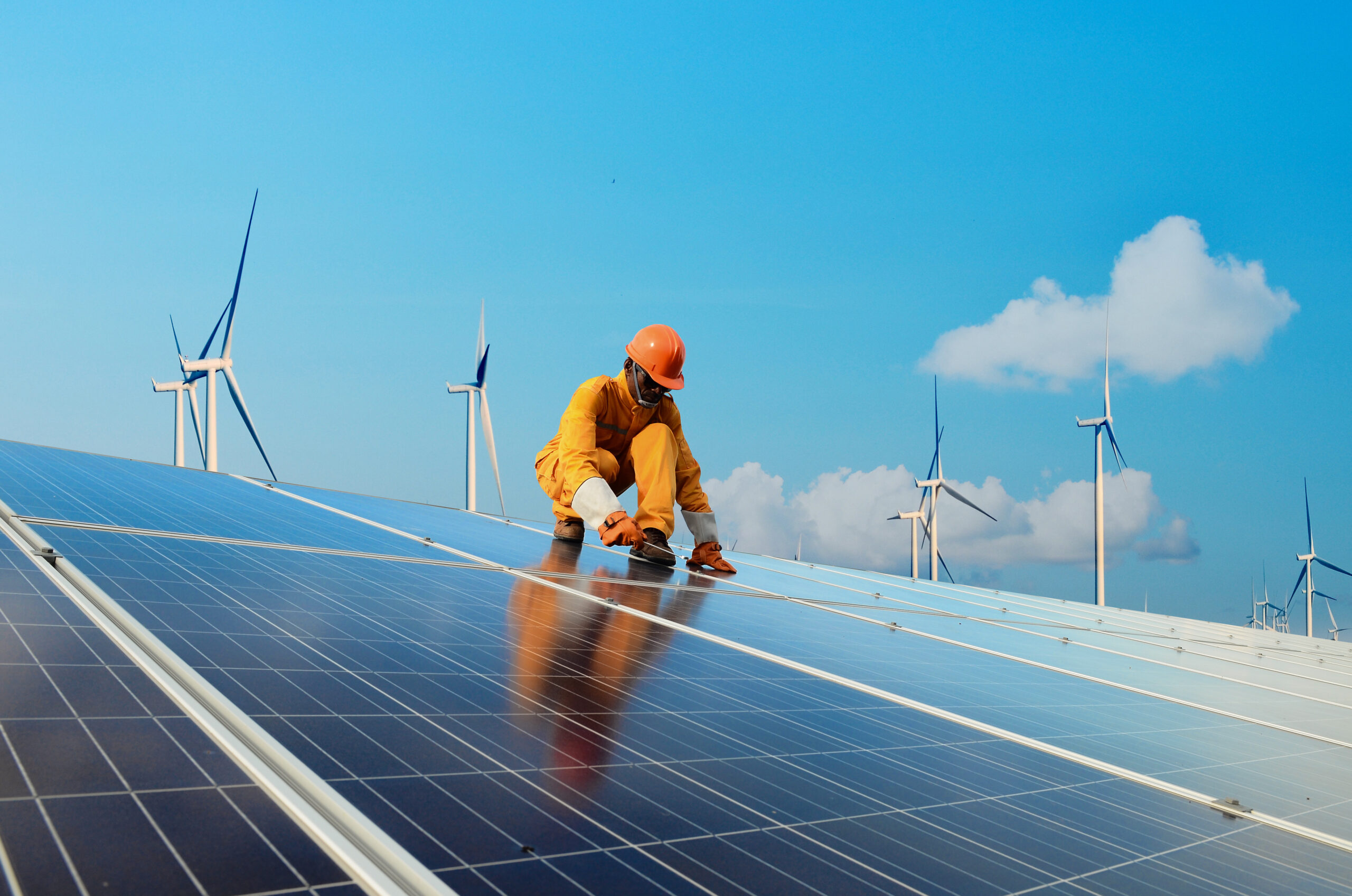 A depiction of clean renewable energy jobs. A person in protective work attre, rain gear and a hardhat and gloves, crouches atop a wide array of solar panels, working on installing or adjusting the clean energy technology. The person appears to be Black. In the background is a blue sky with puffy white clouds. On the horizong are half a dozen large scale wind generators.