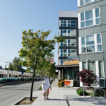 Photo of apartments on a sunny day while a ground-level train comes in. A person in white clothing is centered in the center on the sidewalk