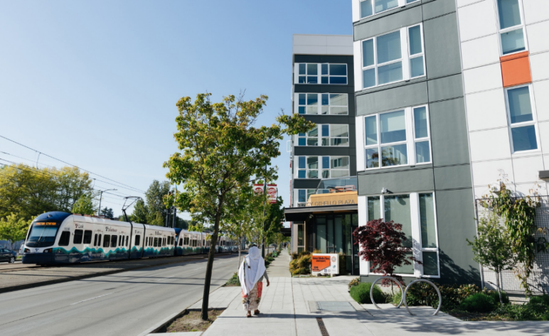 Photo of apartments on a sunny day while a ground-level train comes in. A person in white clothing is centered in the center on the sidewalk