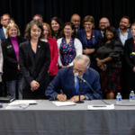 a group of people look on smiling behind a woman as she stands behind a man signing a formal document