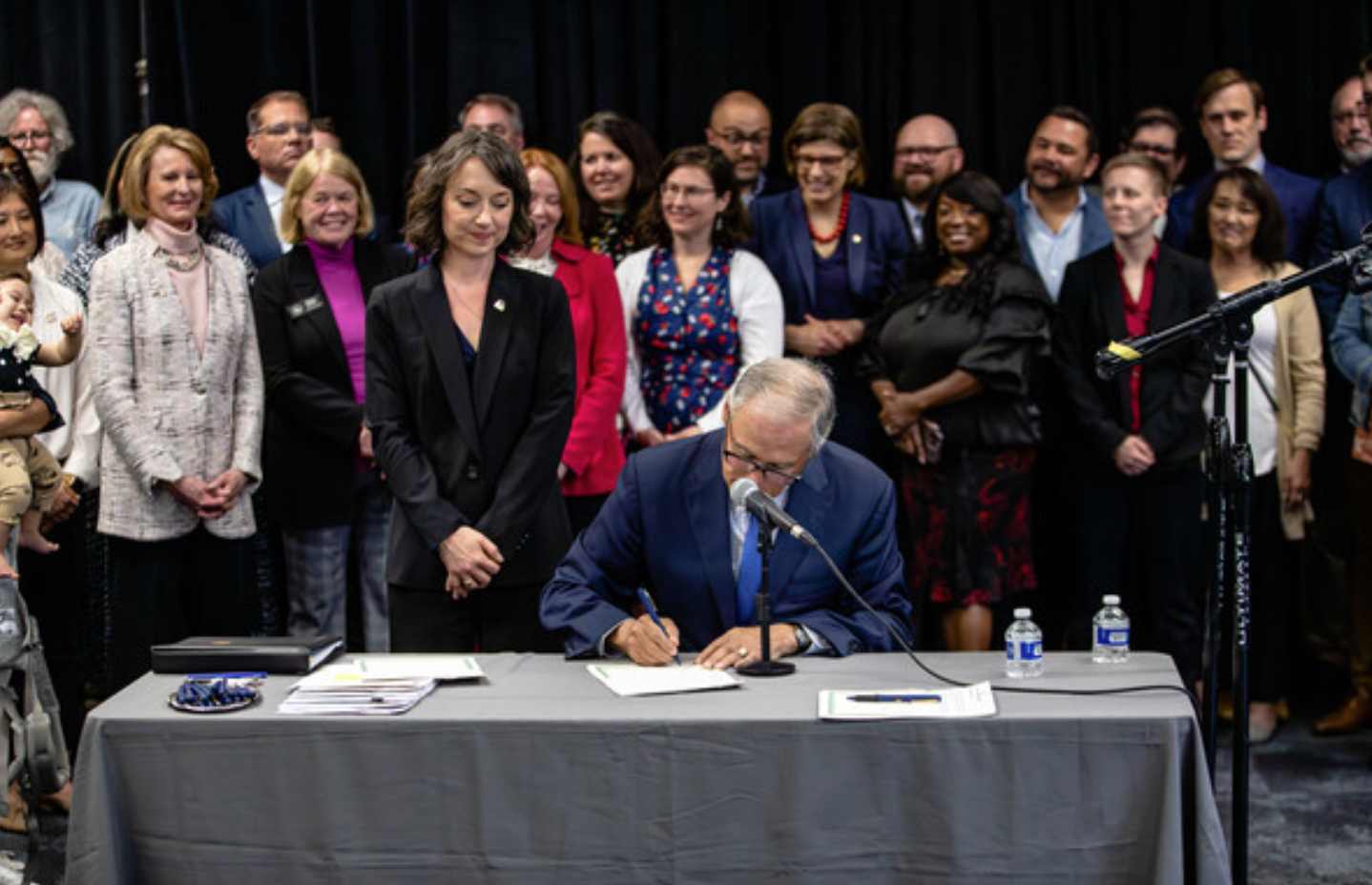 a group of people look on smiling behind a woman as she stands behind a man signing a formal document