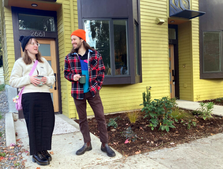 A casually dressed couple in their 20s, one woman and one man, stand in front of a mustard-yellow townhouse with the neighbors' door to the same building visible nearby.