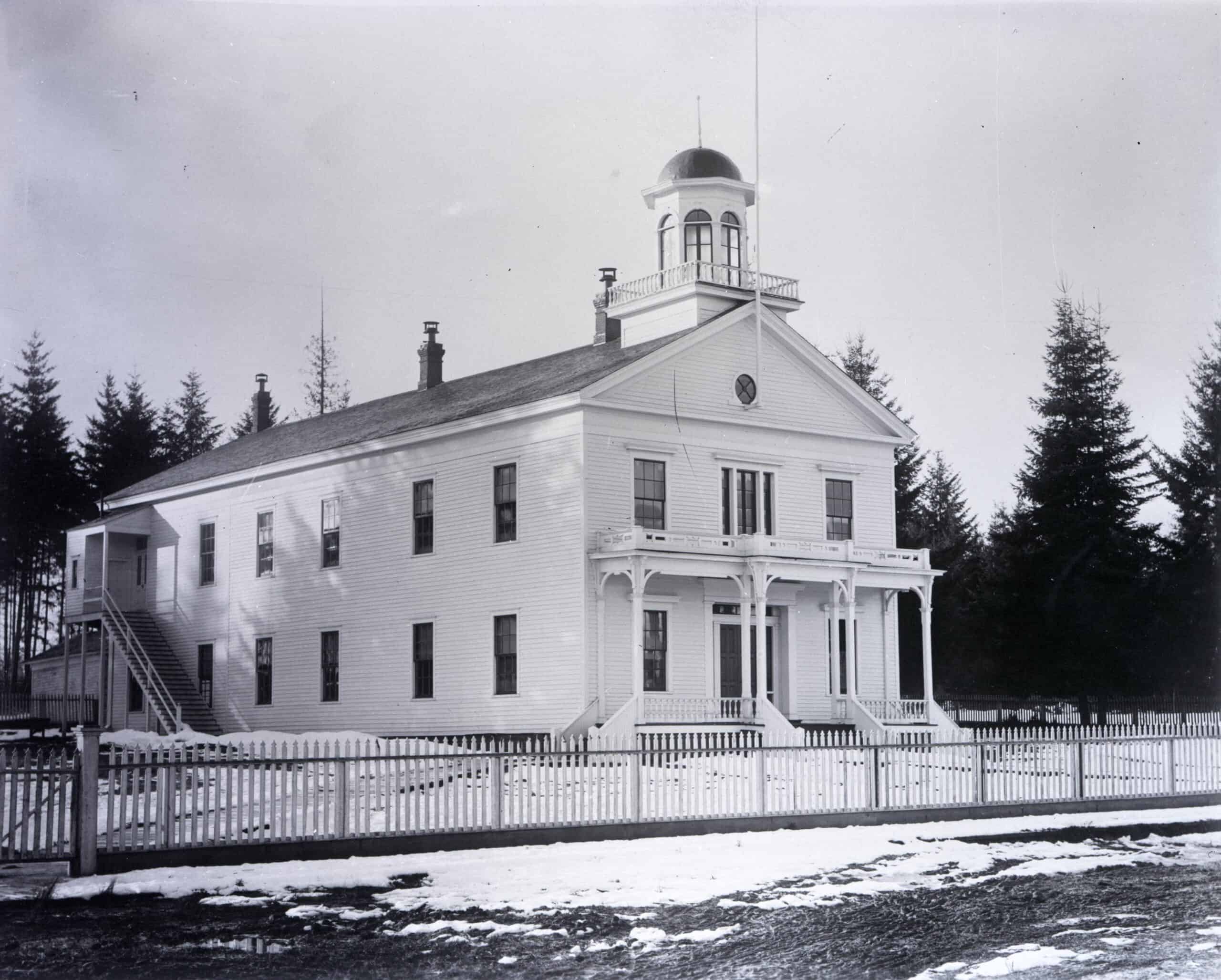 Image shows a two story clapboard building with a cupola on top. Sashed windows are visible along the front and right sides of the building. The front of the building includes a wide porch, and the second story has a widows walk in front. A flagpole rises from the uppermost story. A rear addition, which was added in 1889, is visible on the right side of the building. Snow is on the ground, and evergreen trees stand behind the building. A white picket fence wraps around the building.